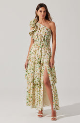 A woman is wearing a long, floral-print dress in shades of cream, green, and peach. The dress features a ruffled detail on one shoulder, an elastic waistband, and a high slit on the front left side. She is accessorized with gold earrings and is wearing strappy white high-heeled sandals.